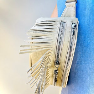 Puede incluir: Bolso bandolera de cuero color crema con flecos. El bolso tiene dos cremalleras plateadas y una correa ajustable. El bolso se lleva sobre el hombro, mostrando su dise&ntilde;o y textura. Medidas en cent&iacute;metros.