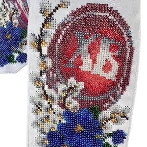 May include: Close-up of white fabric with detailed beadwork. A large red and white beaded emblem is surrounded by white and gold floral accents, and blue beaded flowers. The design is intricate and ornate, showcasing a high level of craftsmanship.