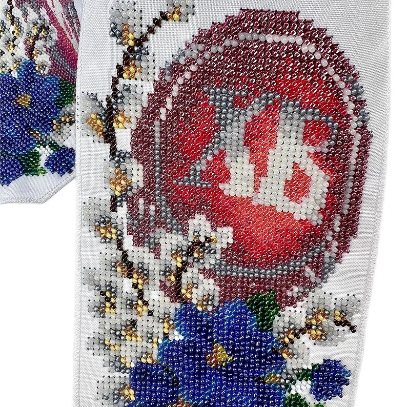 May include: Close-up of a white fabric with intricate beadwork. The design features a large red and white beaded emblem, surrounded by white and gold floral accents, and blue beaded flowers. The overall aesthetic is detailed and ornate.