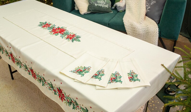 May include: A white tablecloth with a red and green holly and poinsettia design. The tablecloth is set with a matching runner and five napkins.