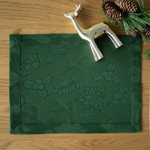 Op de afbeelding: Een donkergroene rechthoekige placemat met een reli&euml;fpatroon van hulst, bessen en dennennaalden. Een goudkleurig hertenbeeldje ligt op de placemat, met dennenappels en groen in de achtergrond.