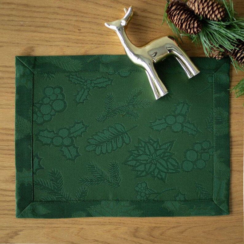 Op de afbeelding: Een donkergroene rechthoekige placemat met een reli&euml;fpatroon van hulst, bessen en dennennaalden. Een goudkleurig hertenbeeldje ligt op de placemat, met dennenappels en groen in de achtergrond.