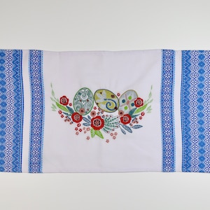 May include: White Easter table runner with blue geometric patterned borders and fringed edges. The center features an embroidered design of three decorated Easter eggs surrounded by red flowers and green leaves.