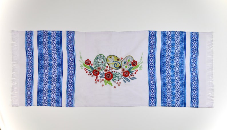 May include: White Easter table runner with blue geometric patterned borders and fringed edges. The center features an embroidered design of three decorated Easter eggs surrounded by red flowers and green leaves.