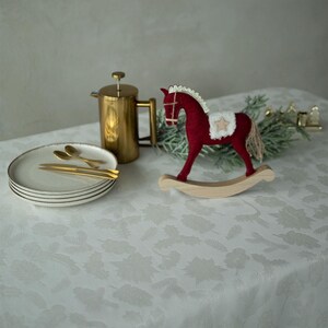 Op de afbeelding: Een feestelijke setting met een rood schommelpaard, een gouden cafeti&egrave;re en borden met gouden bestek. De items zijn geplaatst op een witte tafelkleed met een subtiel bloemmotief, wat een gezellige kerstsfeer cre&euml;ert.