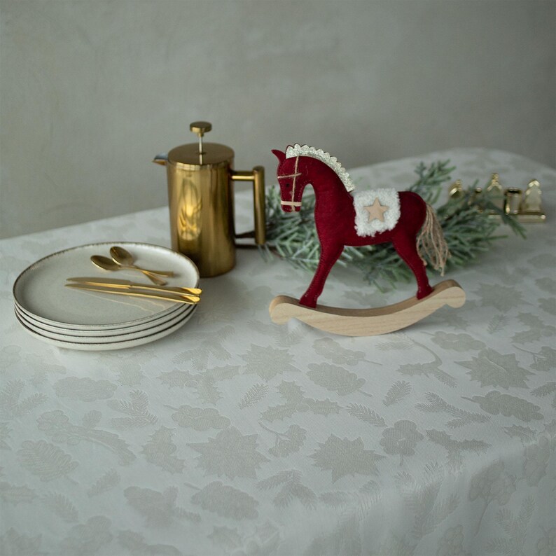 Op de afbeelding: Een feestelijke setting met een rood schommelpaard, een gouden cafeti&egrave;re en borden met gouden bestek. De items zijn geplaatst op een witte tafelkleed met een subtiel bloemmotief, wat een gezellige kerstsfeer cre&euml;ert.