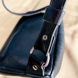 May include: A black leather shoulder bag with a silver metal ring and a strap. The bag is closed and the strap is folded over the top of the bag.