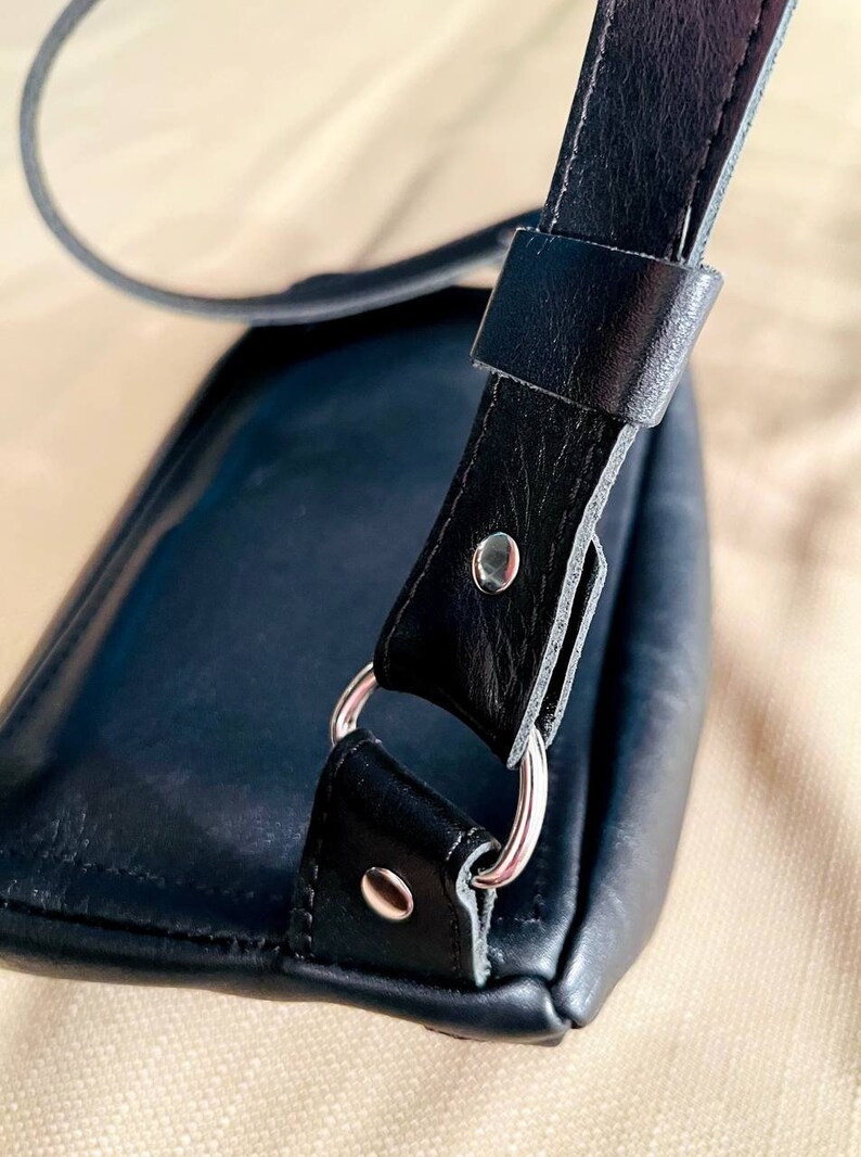 May include: A black leather shoulder bag with a silver metal ring and a strap. The bag is closed and the strap is folded over the top of the bag.