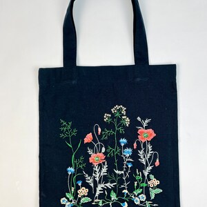 以下が含まれることがあります： 黒色のトートバッグ、花の刺繍デザイン。 刺繍は赤いポピー、青いヤグルマギク、その他のワイルドフラワーと緑の茎と葉が特徴です。 バッグには黒いハンドルが2つ付いています。