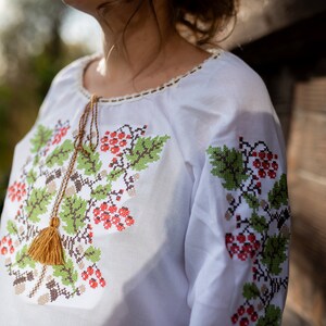 May include: White embroidered blouse with a floral pattern featuring red berries and green leaves. The blouse has a tassel detail at the neckline.
