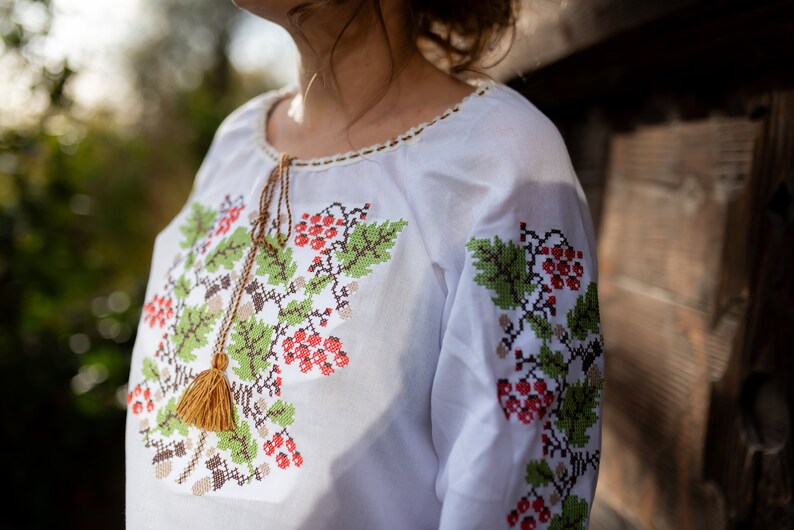 May include: White embroidered blouse with a floral pattern featuring red berries and green leaves. The blouse has a tassel detail at the neckline.