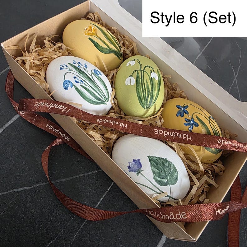May include: A set of five decorative Easter eggs in a cardboard box with a clear lid. Each egg is painted with a different floral design in shades of yellow, white, and green. A brown ribbon with "Handmade" text is wrapped around the box.