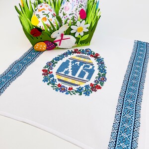 Peut inclure: Serviette de cuisine blanche brod&eacute;e avec une bordure bleue et blanche. La serviette pr&eacute;sente un motif de point de croix d'un &oelig;uf de P&acirc;ques bleu et jaune entour&eacute; d'une couronne de fleurs.