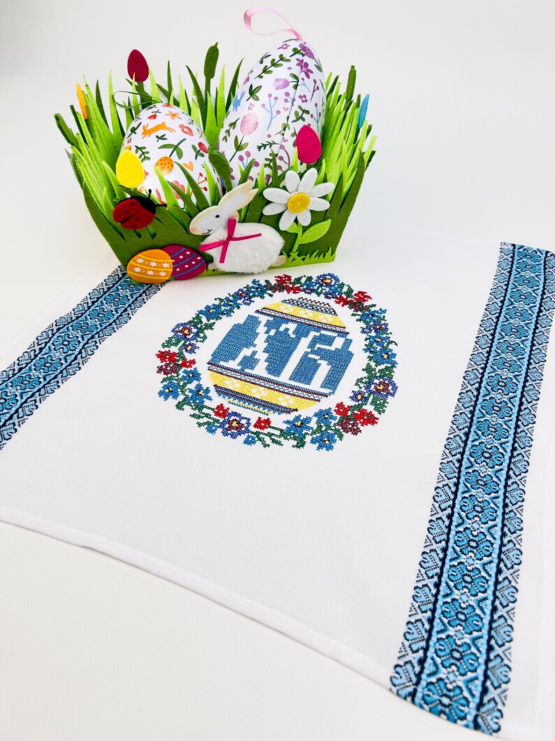 Peut inclure: Serviette de cuisine blanche brod&eacute;e avec une bordure bleue et blanche. La serviette pr&eacute;sente un motif de point de croix d'un &oelig;uf de P&acirc;ques bleu et jaune entour&eacute; d'une couronne de fleurs.