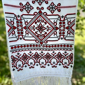 Puede incluir: Textil blanco con intrincado bordado de punto de cruz en rojo y negro. El dise&ntilde;o presenta patrones geom&eacute;tricos, incluyendo diamantes y motivos florales, con un borde con flecos. El bordado est&aacute; centrado en la tela, creando un dise&ntilde;o sim&eacute;trico.
