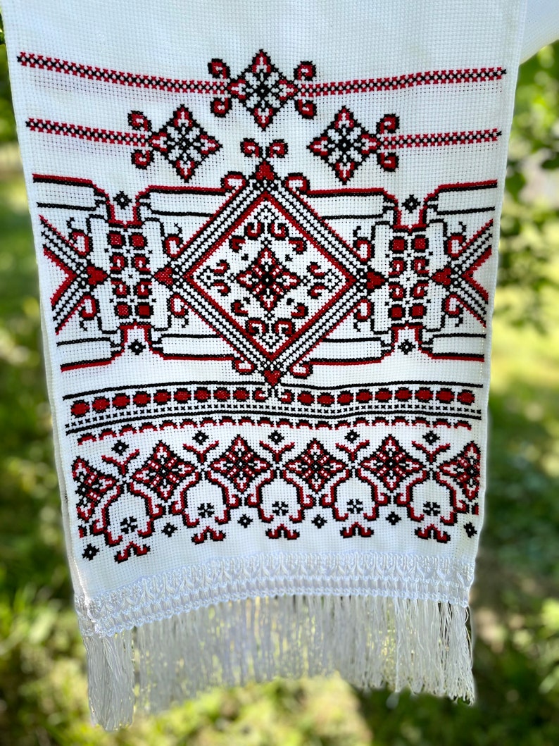 Puede incluir: Textil blanco con intrincado bordado de punto de cruz en rojo y negro. El dise&ntilde;o presenta patrones geom&eacute;tricos, incluyendo diamantes y motivos florales, con un borde con flecos. El bordado est&aacute; centrado en la tela, creando un dise&ntilde;o sim&eacute;trico.