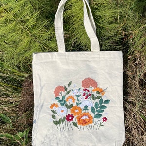 May include: A white canvas tote bag with an embroidered floral design. The flowers are in shades of orange, red, and pink.