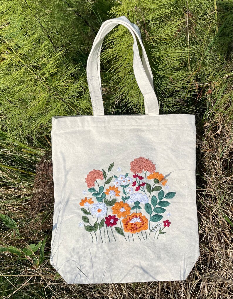 May include: A white canvas tote bag with an embroidered floral design. The flowers are in shades of orange, red, and pink.