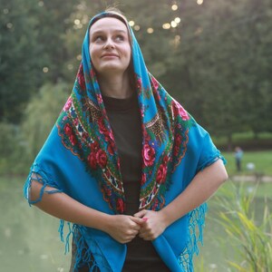 May include: A vibrant blue shawl with a floral pattern in red, pink, and green. The shawl has a fringed edge and is draped over the head and shoulders. The background is a natural outdoor setting.