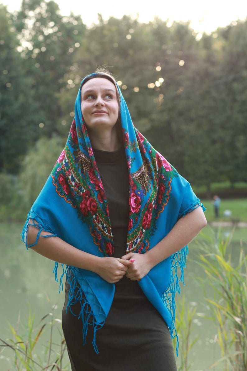 May include: A vibrant blue shawl with a floral pattern in red, pink, and green. The shawl has a fringed edge and is draped over the head and shoulders. The background is a natural outdoor setting.