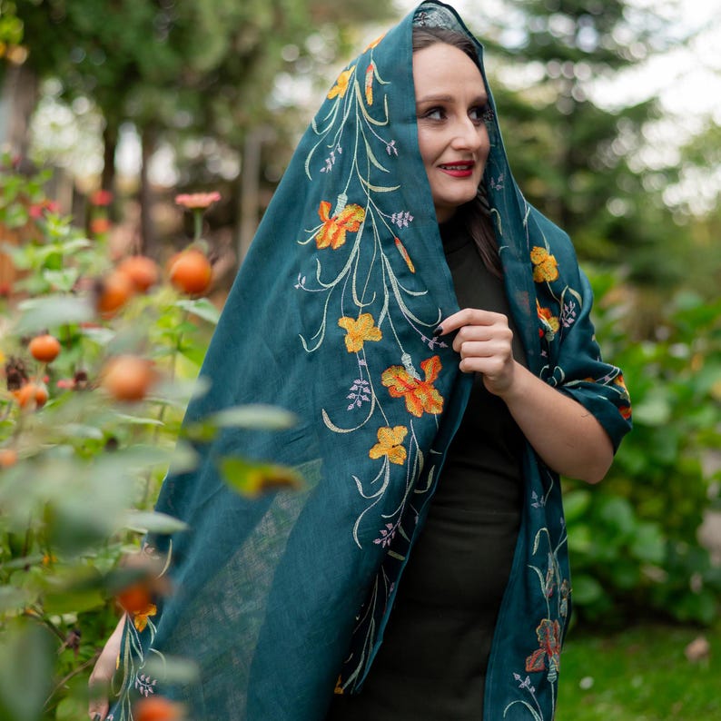 May include: Teal-colored shawl with embroidered floral design in yellow and orange. The shawl is draped over the head and shoulders, creating a hooded effect. The floral embroidery features detailed petals and stems, adding a touch of elegance to the garment.