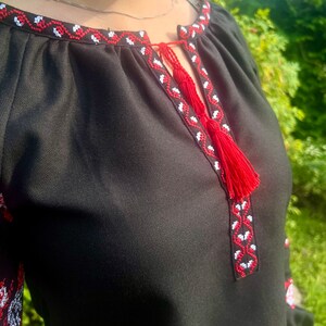 K&ouml;nnte beinhalten: Schwarze bestickte Bluse mit rotem und wei&szlig;em Besatz. Der Ausschnitt hat ein Quasten-Detail.