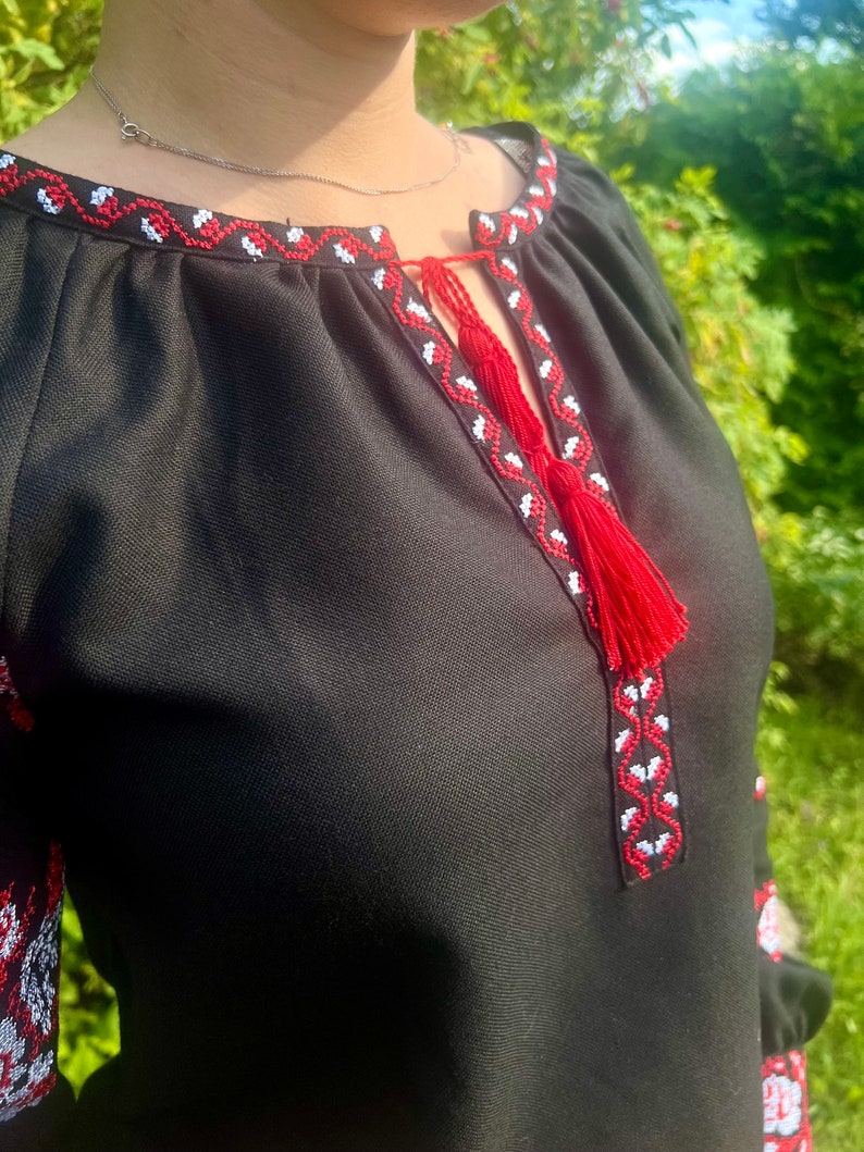 K&ouml;nnte beinhalten: Schwarze bestickte Bluse mit rotem und wei&szlig;em Besatz. Der Ausschnitt hat ein Quasten-Detail.