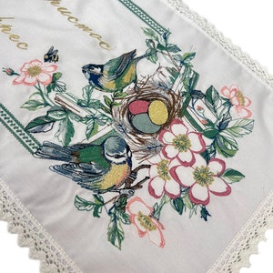 以下が含まれることがあります： 鳥、花、卵の巣が刺繍された白いテーブルランナー。 ランナーは緑色の縁取りと繊細なレースの縁取りが施されています。 金色の文字が左上隅に見えます。 デザインはピンク、緑、青、黄色の色を使用しています。