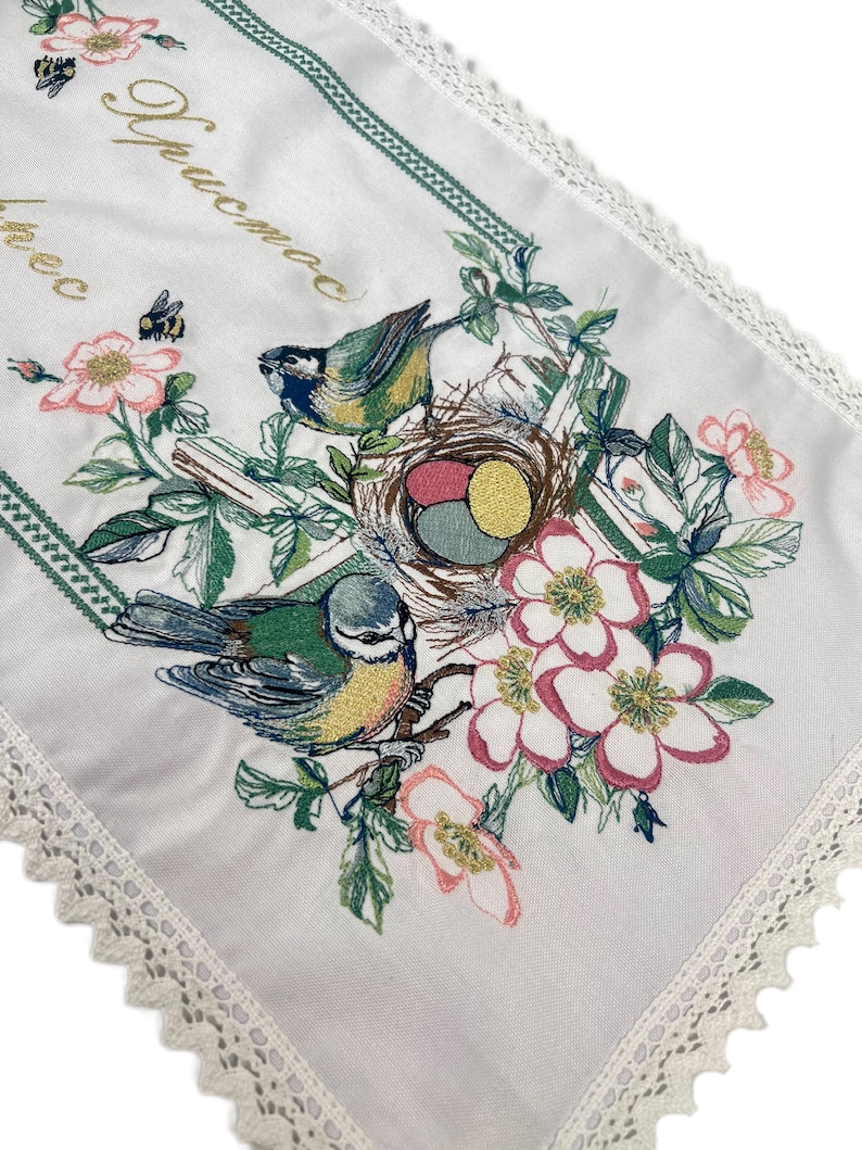 以下が含まれることがあります： 鳥、花、卵の巣が刺繍された白いテーブルランナー。 ランナーは緑色の縁取りと繊細なレースの縁取りが施されています。 金色の文字が左上隅に見えます。 デザインはピンク、緑、青、黄色の色を使用しています。