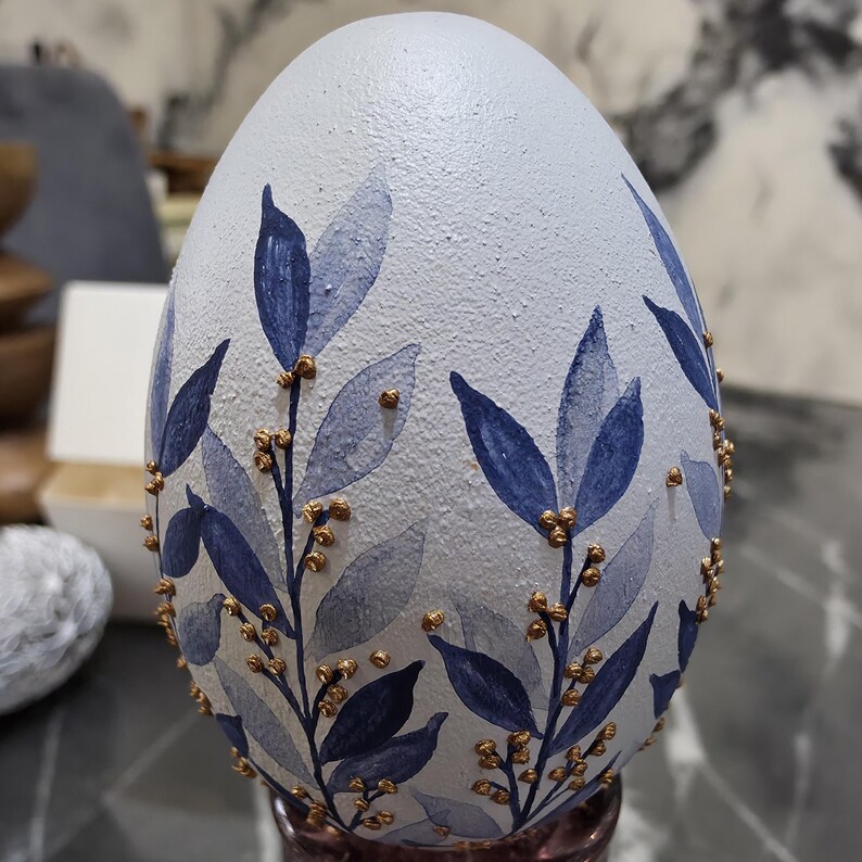 Puede incluir: Un huevo decorativo con una superficie texturizada azul claro. El huevo est&aacute; adornado con hojas azules estilo acuarela y detalles dorados, creando un dise&ntilde;o elegante inspirado en la naturaleza. El huevo se exhibe sobre un soporte rosa.