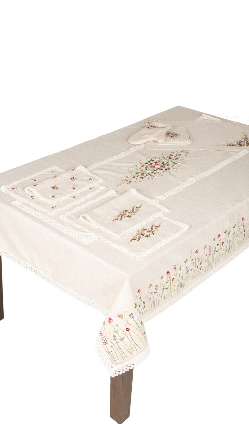May include: A white tablecloth with embroidered floral designs and a lace trim. The tablecloth is set with four placemats and a set of pot holders.