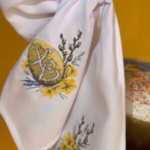 K&ouml;nnte beinhalten: Wei&szlig;e bestickte Stoffserviette mit einem gelben gestickten Ei mit den kyrillischen Buchstaben "XB" und einer gelben bestickten Glocke mit einem Zweig gelber Blumen.