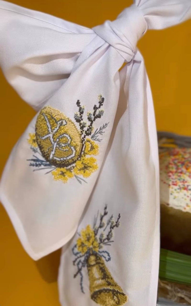 K&ouml;nnte beinhalten: Wei&szlig;e bestickte Stoffserviette mit einem gelben gestickten Ei mit den kyrillischen Buchstaben "XB" und einer gelben bestickten Glocke mit einem Zweig gelber Blumen.