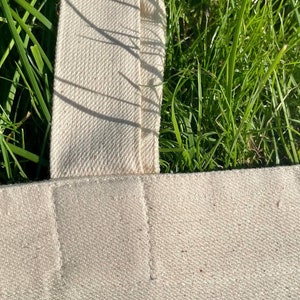 May include: Close-up of a white canvas tote bag with a thick, white canvas strap. The strap is sewn to the bag with a straight stitch.