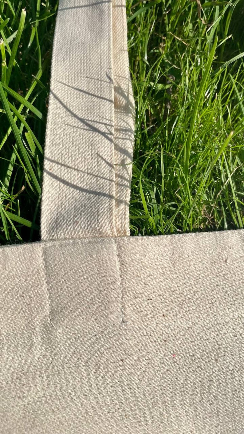 May include: Close-up of a white canvas tote bag with a thick, white canvas strap. The strap is sewn to the bag with a straight stitch.