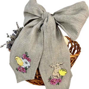 May include: A grey linen bow featuring embroidered Easter designs. The embroidery depicts a yellow chick and a rabbit with a yellow egg, surrounded by pink flowers. The bow is positioned over a woven basket.