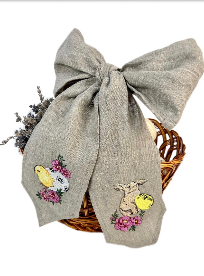May include: A grey linen bow featuring embroidered Easter designs. The embroidery depicts a yellow chick and a rabbit with a yellow egg, surrounded by pink flowers. The bow is positioned over a woven basket.