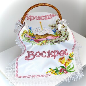 May include: A wicker basket with a white embroidered cloth. The cloth features the word "Воскрес" in red, an embroidered cake, and floral designs. It has a lace trim and a woven handle.