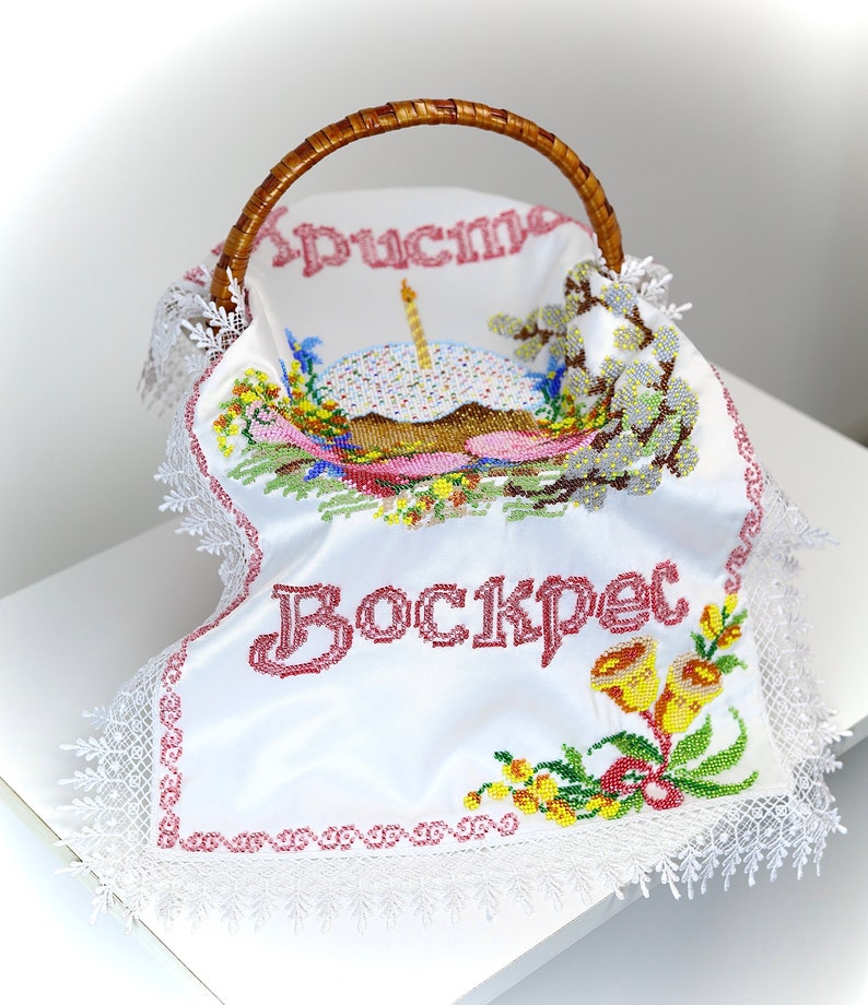 May include: A wicker basket with a white embroidered cloth. The cloth features the word "Воскрес" in red, an embroidered cake, and floral designs. It has a lace trim and a woven handle.