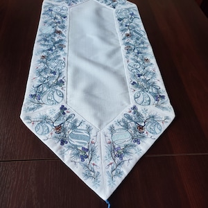 Puede incluir: Un camino de mesa blanco con un diseño bordado en azul y verde de ramas de pino, bayas y adornos. El camino de mesa tiene borlas azules en cada extremo.