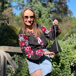 May include: A woman wearing a black embroidered blouse with red and white floral designs, blue denim shorts, and sunglasses. She is holding a black triangular handbag.