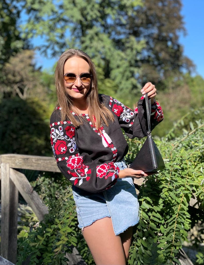 May include: A woman wearing a black embroidered blouse with red and white floral designs, blue denim shorts, and sunglasses. She is holding a black triangular handbag.
