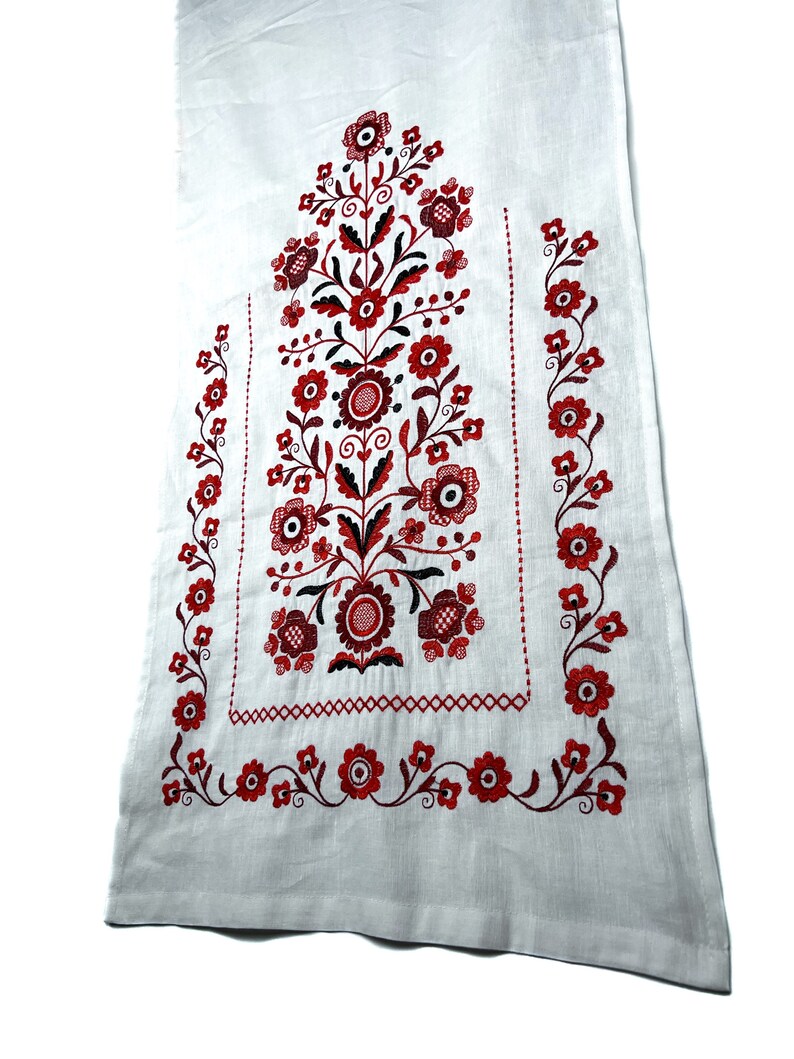 K&ouml;nnte beinhalten: Wei&szlig;er Leinenstoff mit roter und schwarzer Stickerei in Blumenform. Das Design zeigt ein zentrales Blumenmotiv mit kleineren Blumenmotiven an den R&auml;ndern.