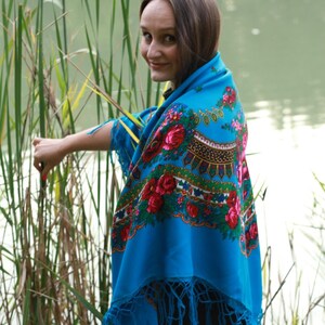 May include: A vibrant blue shawl with a floral pattern featuring red and pink roses, green leaves, and gold accents. The shawl has a fringed edge. The image is set outdoors near tall grass and water.
