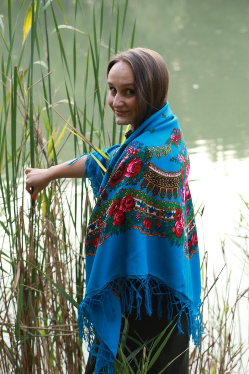 May include: A vibrant blue shawl with a floral pattern featuring red and pink roses, green leaves, and gold accents. The shawl has a fringed edge. The image is set outdoors near tall grass and water.