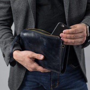 May include: A dark blue leather clutch bag with a zipper closure and a gold metal zipper pull. A black smartphone is partially visible inside the bag.