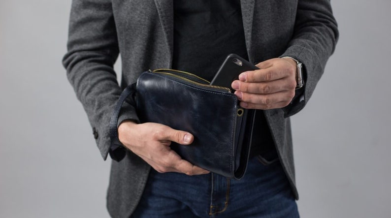 May include: A dark blue leather clutch bag with a zipper closure and a gold metal zipper pull. A black smartphone is partially visible inside the bag.