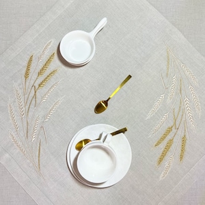 Peut inclure: Une nappe beige avec des &eacute;pis de bl&eacute; brod&eacute;s en or et cr&egrave;me. La table est dress&eacute;e avec des bols, des assiettes et une tasse &agrave; th&eacute; en c&eacute;ramique blanche, ainsi que des cuill&egrave;res dor&eacute;es, cr&eacute;ant une sc&egrave;ne de repas &eacute;l&eacute;gante.