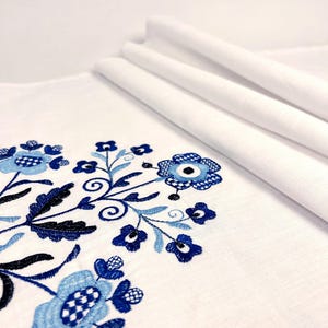 May include: A white fabric textile featuring intricate embroidery in shades of navy, royal, and light blue. The design consists of stylized floral motifs with circular centers, delicate petals, and swirling stems arranged in a symmetrical pattern.