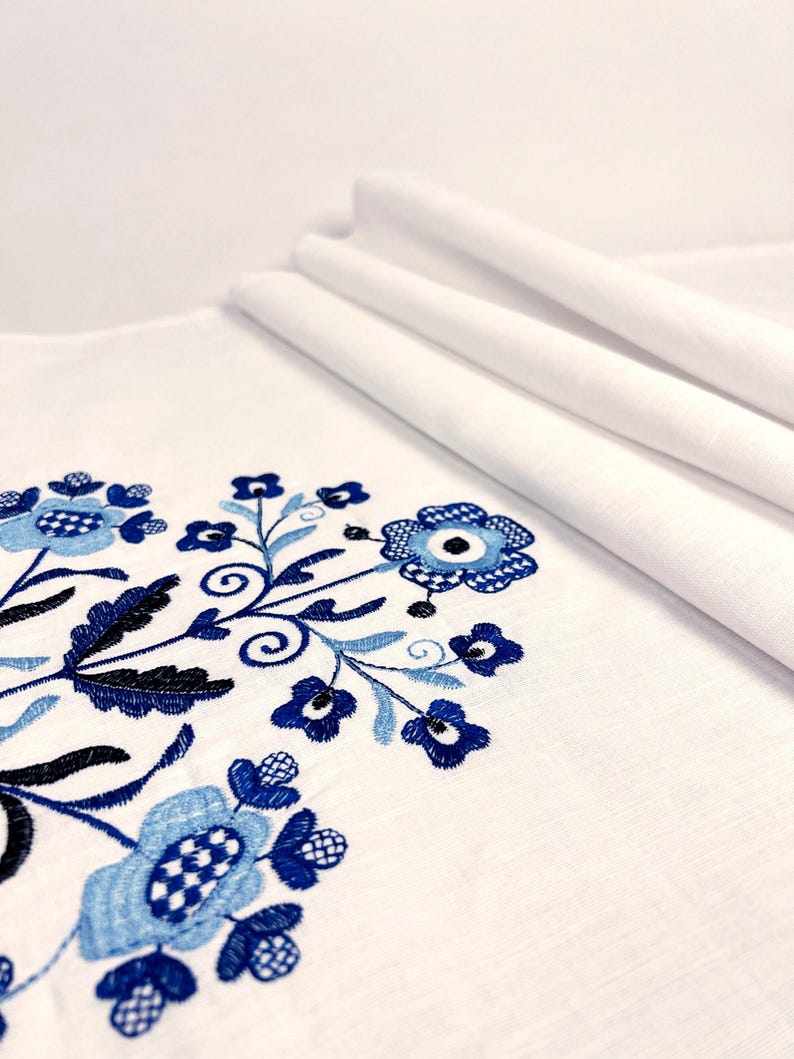 May include: A white fabric textile featuring intricate embroidery in shades of navy, royal, and light blue. The design consists of stylized floral motifs with circular centers, delicate petals, and swirling stems arranged in a symmetrical pattern.